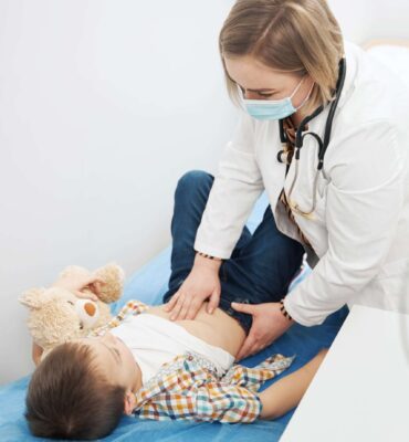 Little boy lying on hospital daybed and holding toy while woman pediatrician doing abdominal palpation