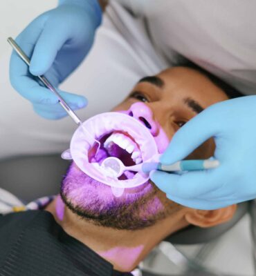 Young man with patient bib on a dental chair, pink light oriented on his teeth and a dentist who sits next to him wearing blue medical gloves while examining oral cavity